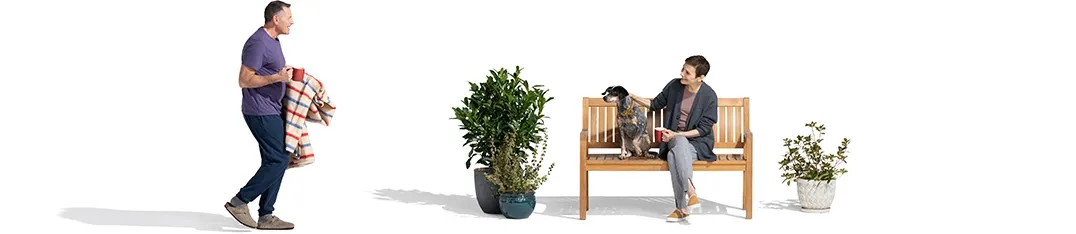 Between a pair of potted plants, a woman sits on a wood bench with her dog. She watches as her husband approaches with a Christmas-colored blanket and a red mug.